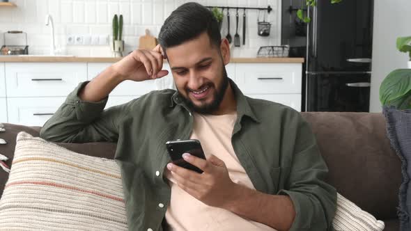 Positive Indian or Arabian Attractive Guy Using His Smartphone While Sits on Sofa in Living Room