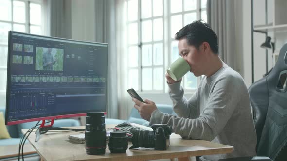 Asian Video Editor Man Using Mobile Phone And Drinking Coffee While Using Computer For Editing Video