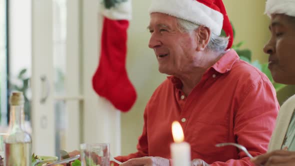Diverse senior man and woman in santa hats listening and laughing at christmas dinner table at home