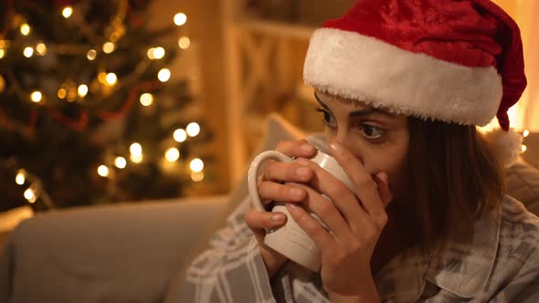 Woman in Pajama and Red Santa Hat Sitting on Couch Watching TV and Drinking Hot Chocolate From Mug