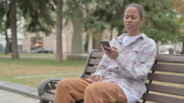 African Woman Reacting to Loss on Smartphone While Sitting Outdoor on Bench