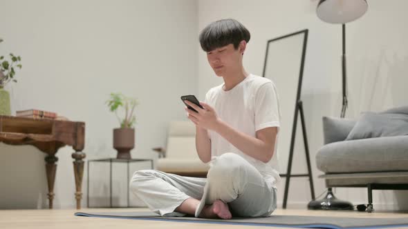 Young Asian Man Using Smartphone on Yoga Mat at Home