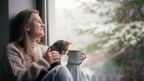A Beautiful Young Girl Sits on the Windowsill with a Cute Cat