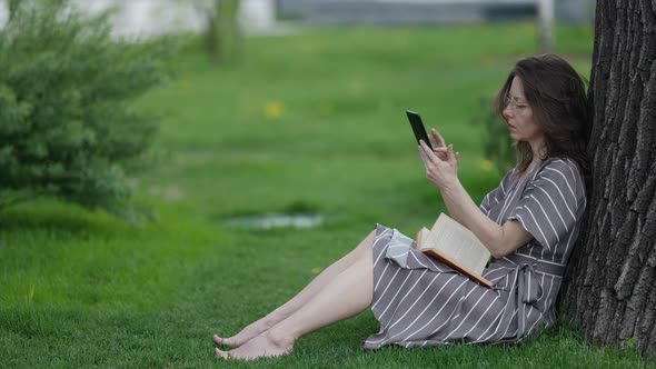 a Brunette in a Striped Dress Glasses and Barefoot is Sitting Under a Tree with a Book on Knees