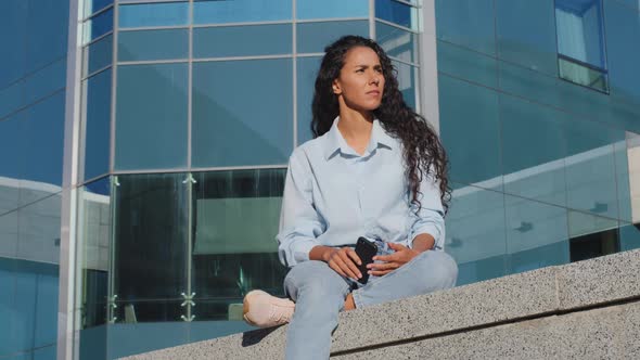 Pensive Stressed Young Woman Girl with Mobile Phone Sits in City Near Building Thinks About Problem