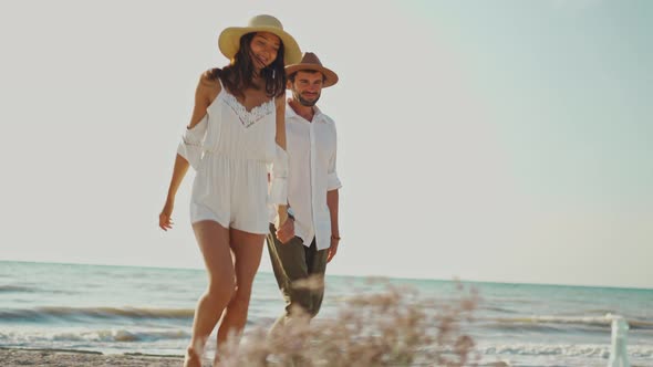 Romantic Couple in Hats Walking Along Sandy Beach Holding Hands