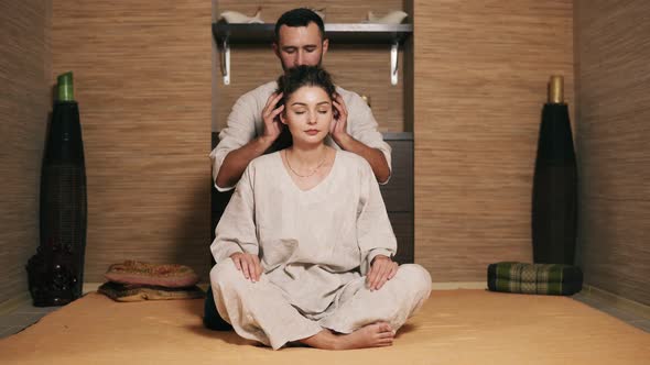 Attractive Thai Massagist is Massaging Young Woman's Head Sitting Behind Her