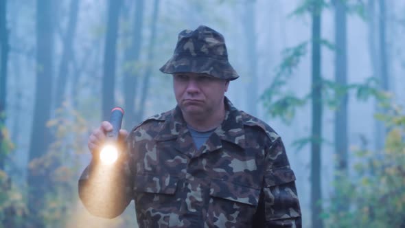 A Man in Camouflage Is Walking Through the Forest with a Flashlight in His Hand