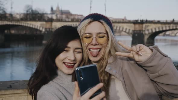 Portrait of Two Girlfriends in Front of Bridge and Embankment of River Holding Smartphone or Mobile