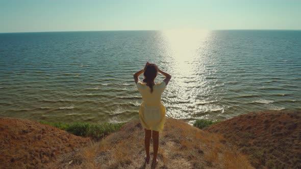 Back View Happy Woman in Yellow Dress Walks on Edge of Clay Hill Over Sea with Morning Sunlight