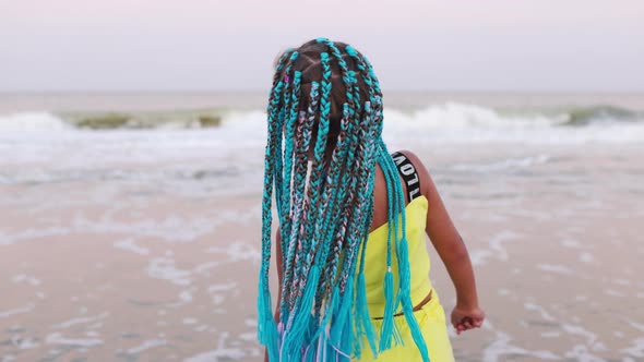 A Girl in a Summer Suit with African Braids Looks at the Sea Horizon While Standing on the Beach