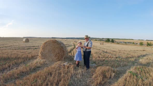 Grandfather and Granddaughter Walking Across the Field with Haystacks. Farmer Grandfather Teaches