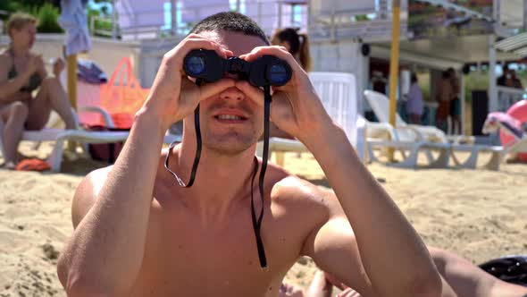 Man on the Beach in Swimming Trunks is Watching Someone Through Binoculars