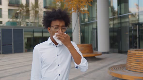 Young African Businessman Walking Outside Office Building and Sneezing in White Napkin.