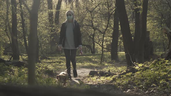 Wide Shot of Young Woman Strolling with Dog in Forest. Blond Caucasian Girl in Face Mask and