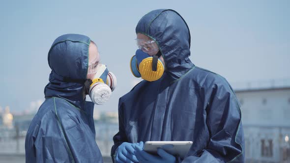 Two Confident Serious People in Antiviral Suits and Respirators Checking Information on Disinfection