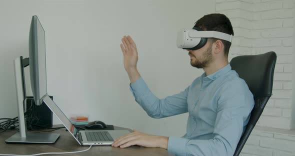 Man in Blue Shirt Sitting at Desk and Using VR Glasses and Laptop for Work