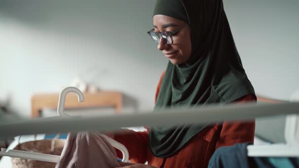 Positive Muslim woman wearing eyeglasses hanging things on a hanger