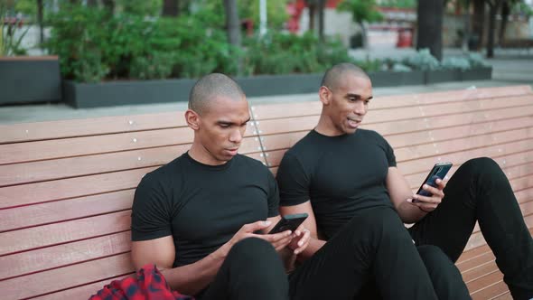 Two positive bald African brothers talking and typing on phone while sitting on the bench