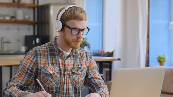 Young Guy in Headphones Studying From Home Using Laptop and Writing Notes