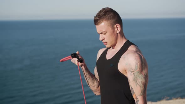 Young Tattooed Man Does Exercises with Elastic Bands at Sunrise with Ocean View