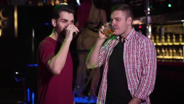 Two Young Caucasian Men Clinking Glasses and Drinking Alcohol in Night Club. Male Friends Resting