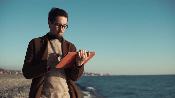 Bearded Man Is Using Flatbed Computer Sitting on Seashore with Panorama of City