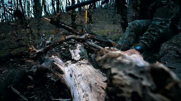 A man with a beard in camouflage with a weapon jumps over a large fallen tree in the forest