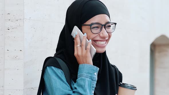 A smiling arabian woman wearing national hijab is talking on her phone while holding a craft cup of