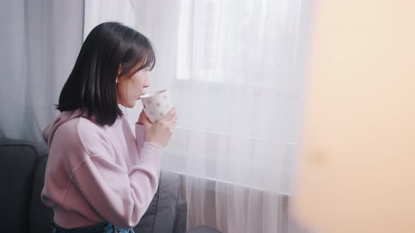 Young Lonely Asian Woman Drinking Morning Coffee and Looking Through the Window