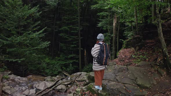 Red Head Girl Tourist with a Backpack, Stands on the Edge of a Cliff of a Waterfall Against a Foggy