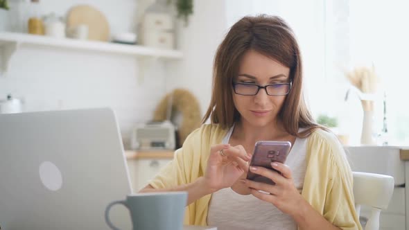 Young Beautiful Female Using Smartphone, Sitting at Table with Laptop at Home