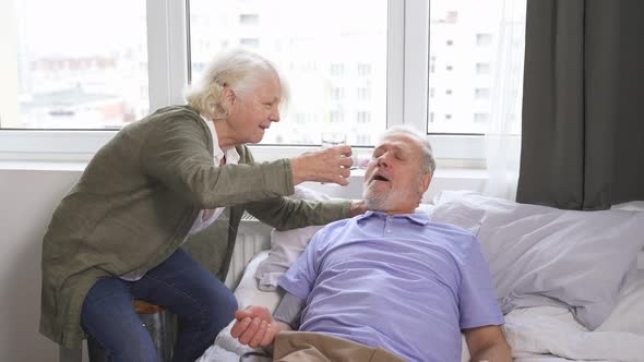 an Elderly Caring Woman Checks Her Husband's Blood Pressure and Gives Her Sick Husband a Glass of