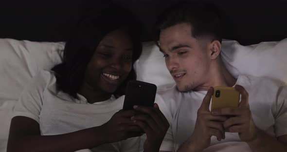 Happy Multi-ethnic Couple in the Bed. They Using Smartphones and Showing Each Other