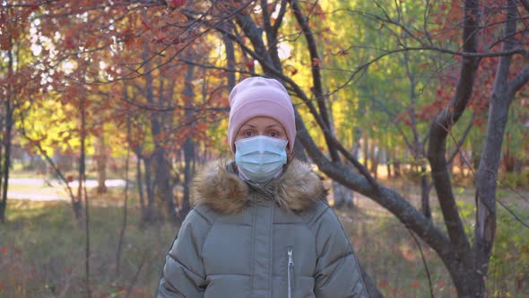 The camera is approaching a girl in a medical mask among autumn trees.