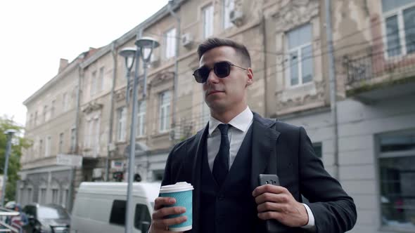 Man in Suit with Sunglasses Halds Cup of Coffee and Corrects Bag Belt on Street