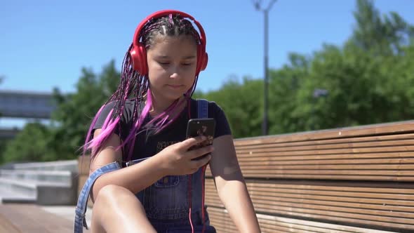 Girl Listening to Music on Headphones Using Smartphone Sitting on a Bench with a Good Mood in Summer