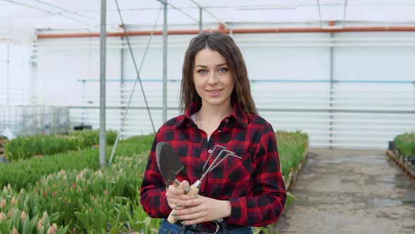 Florist Young Girl in a Redblack Shirt and Jeans Stands on a Background of Rows with Seedlings of