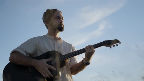 Spanish Bearded Man Playing Guitar Outdoors Against a Blue Sky Lit with Setting Sunlight