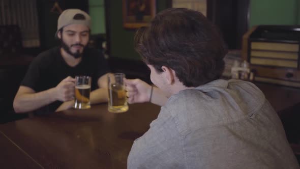 Two Guys Drink Beer Clinking Glasses While Sitting at a Table in a Pub