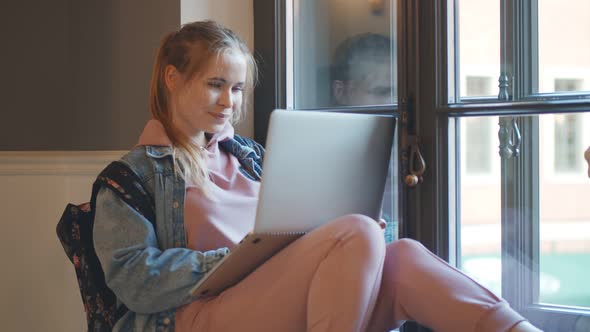 Female Caucasian Student Working on Laptop Sitting on Windowsill in Corridor