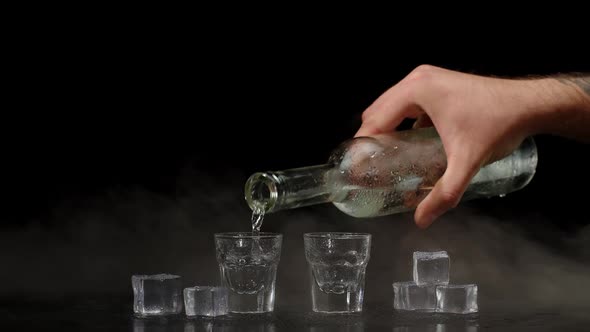 Hand Pours Vodka Tequila or Sake From Bottle Into Shot Glasses on Black Background with Ice Cubes