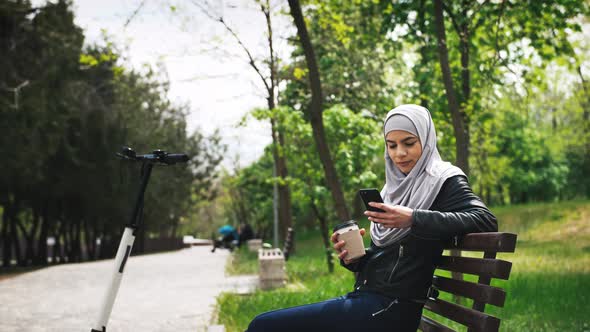 Young Moderm Attractive Muslim Woman Wearing Hijab Sitting in Park and Listening to Music with