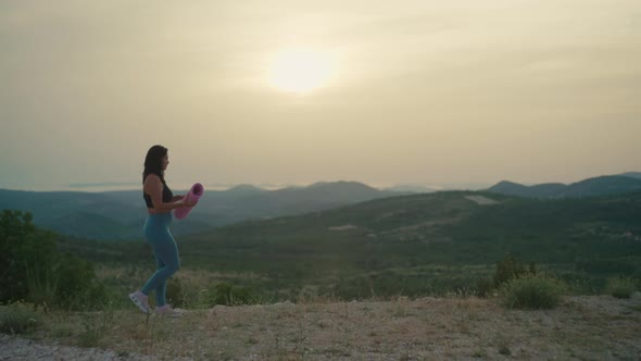 Girl with a Mat Goes to Do Yoga in the Mountains at Sunset