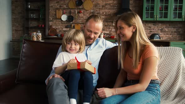 Spouses in Casual Outfit are Smiling Giving Gift Box to Their Child While Sitting on Sofa in Studio