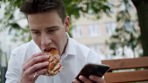 Busy Man Using Smartphone Smiling and Eating a Burger When Sitting on Bench