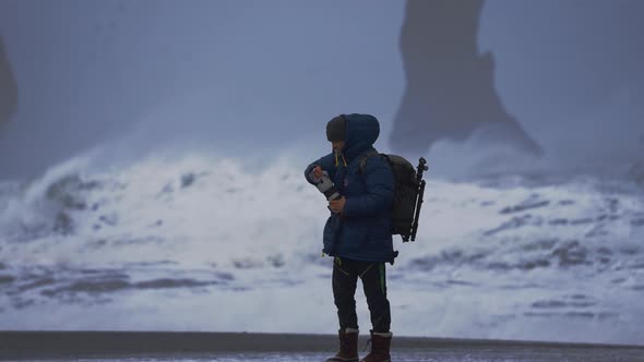 Photographer Cleaning Lens On Black Sand Beach With Stormy Sea