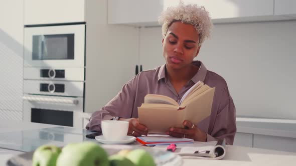 African American Woman Holding Open Book in Hands Looking to Side in Kitchen