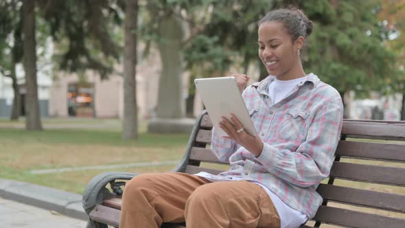 African Woman Celebrating Online Win on Tablet While Sitting Outdoor on Bench