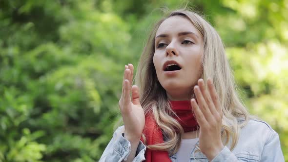 Young Woman Sneezes and Rubs Her Nose Standing on the Park Background